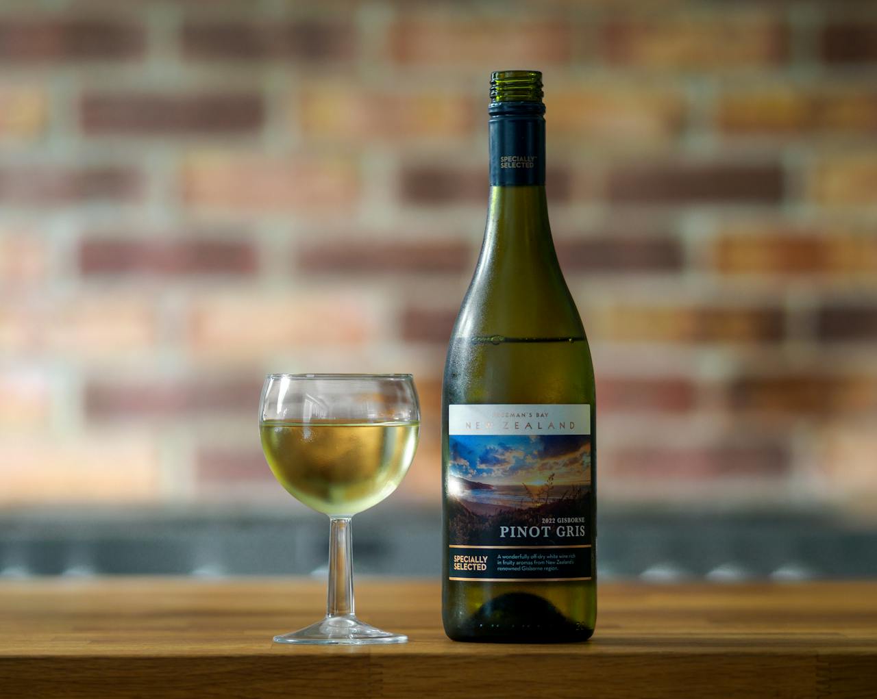 Close-up of a New Zealand Pinot Gris wine bottle and glass set against a blurred brick wall.